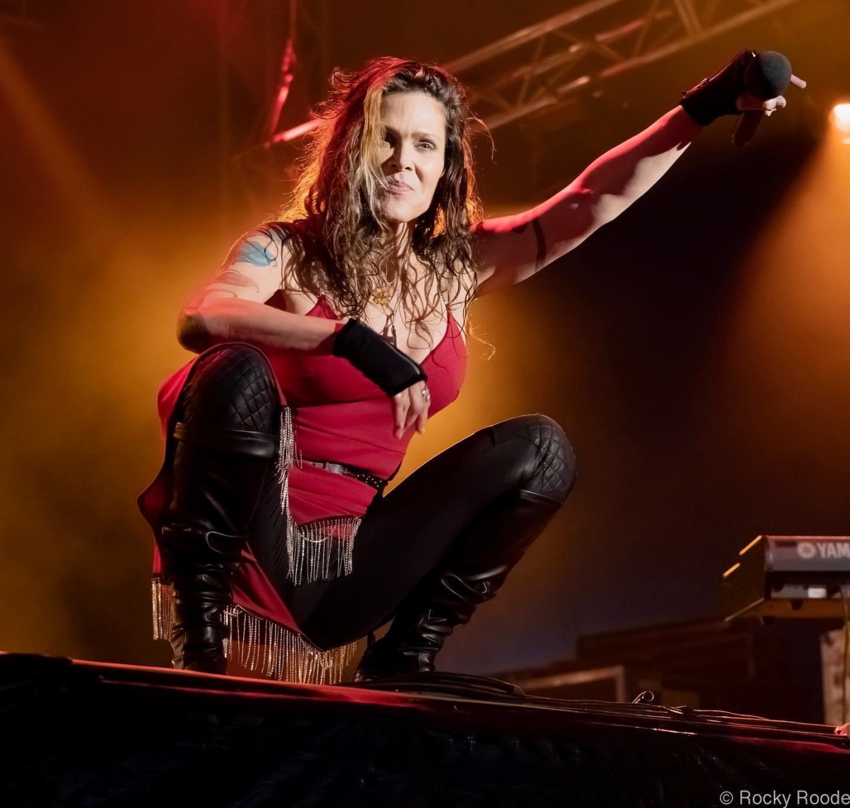 Beth Hart squatting on stage holding her microphone in the air with dramatic lighting behind