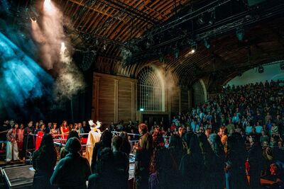 A large audience watches a live performance inside a historic wooden venue. Performers stand on a brightly lit stage in the foreground, surrounded by atmospheric smoke and blue-green lighting, while rows of spectators fill the tiered seating on the right. Exposed timber beams, arched windows, and overhead stage equipment frame the scene.