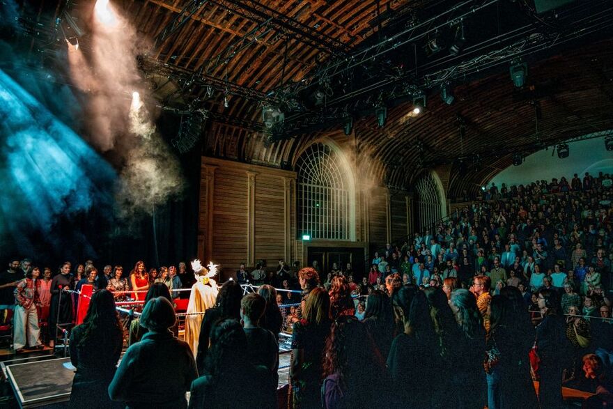 A large audience watches a live performance inside a historic wooden venue. Performers stand on a brightly lit stage in the foreground, surrounded by atmospheric smoke and blue-green lighting, while rows of spectators fill the tiered seating on the right. Exposed timber beams, arched windows, and overhead stage equipment frame the scene.