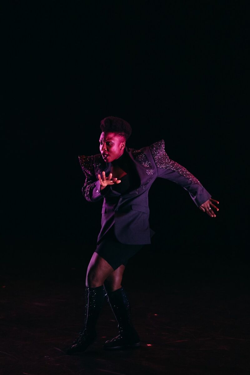 A black performer appears to be dancing, their arms are slightly twisted and their legs appear to be moving. They are wearing an oversized blazer with big pointed shoulder pads.