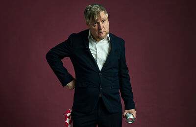 Tim Key wearing a blazer and shirt standing with his hand on his hip, in front of a burgundy background