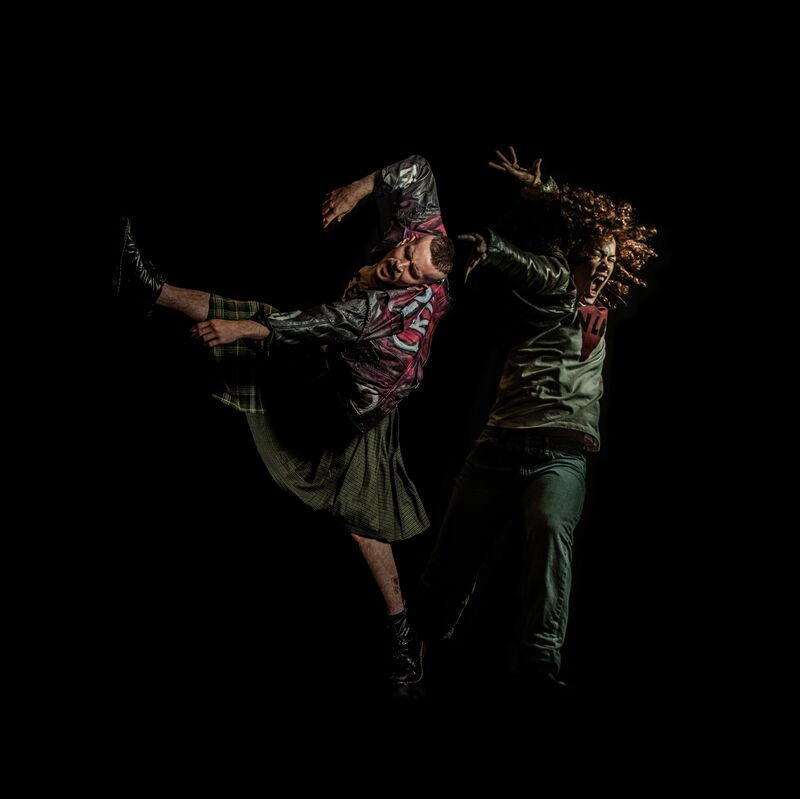 Two dancers jumping, one in a kilt, dark red jacket and short hair kicking one leg to the side and twisting his body. The other in dark clothes with long hair jumping upwards and yelling, both looking extremely angry 
