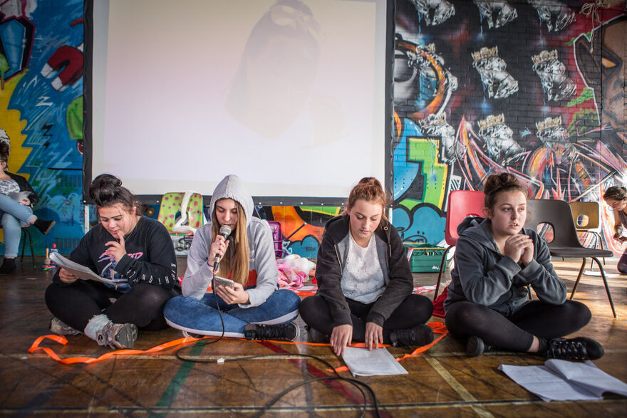 Four young girls sit on the floor, one is holding a script, one a microphone 
