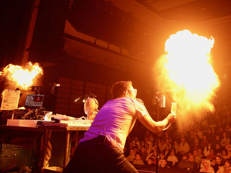 Man hunches over with back to the camera with his arm outstretched towards an audience, holding something exploding with fire