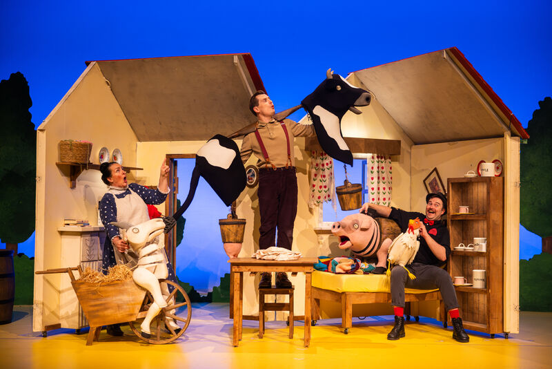 Two men and a woman are in a model house's living room looking excited with puppets of a goat, pig and cow