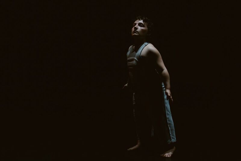 A dancer in high contrast low lighting stands with their face looking upwards