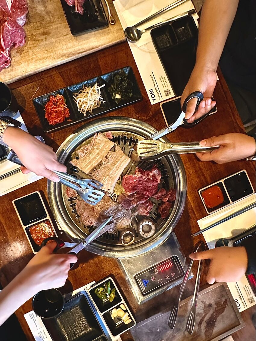 A photo taken above a table, a large hot-plate in the middle with food on, hands reach over the table sharing the food