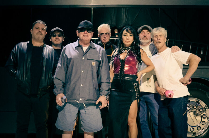 Group of people standing facing the camera. They are wearing shirts and jackets, and one woman is wearing a red top and black long skirt.