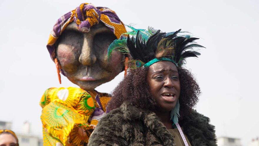 A woman dressed with a head dress stands in front of a life size puppet