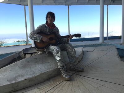 A person wearing a silver spacesuit holding a guitar sitting on a concrete block. Behind them is a big window with a blue sky