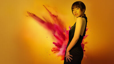 A lady in a black dress over a ellow background with pink powder exploding behind her