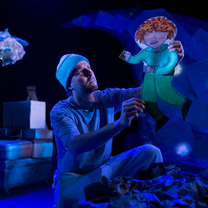 A person in a blue outfit and beanie holds a cut-out of a red-haired character on a dimly lit stage with props.