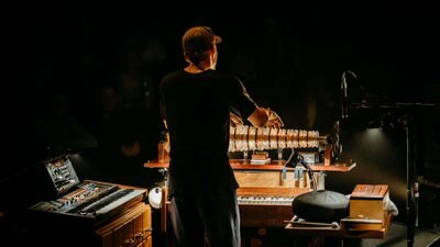 Nils Frahm playing a glass harmonica on stage. Also on stage are a piano, a synth keybord, a hang drum and a microphone