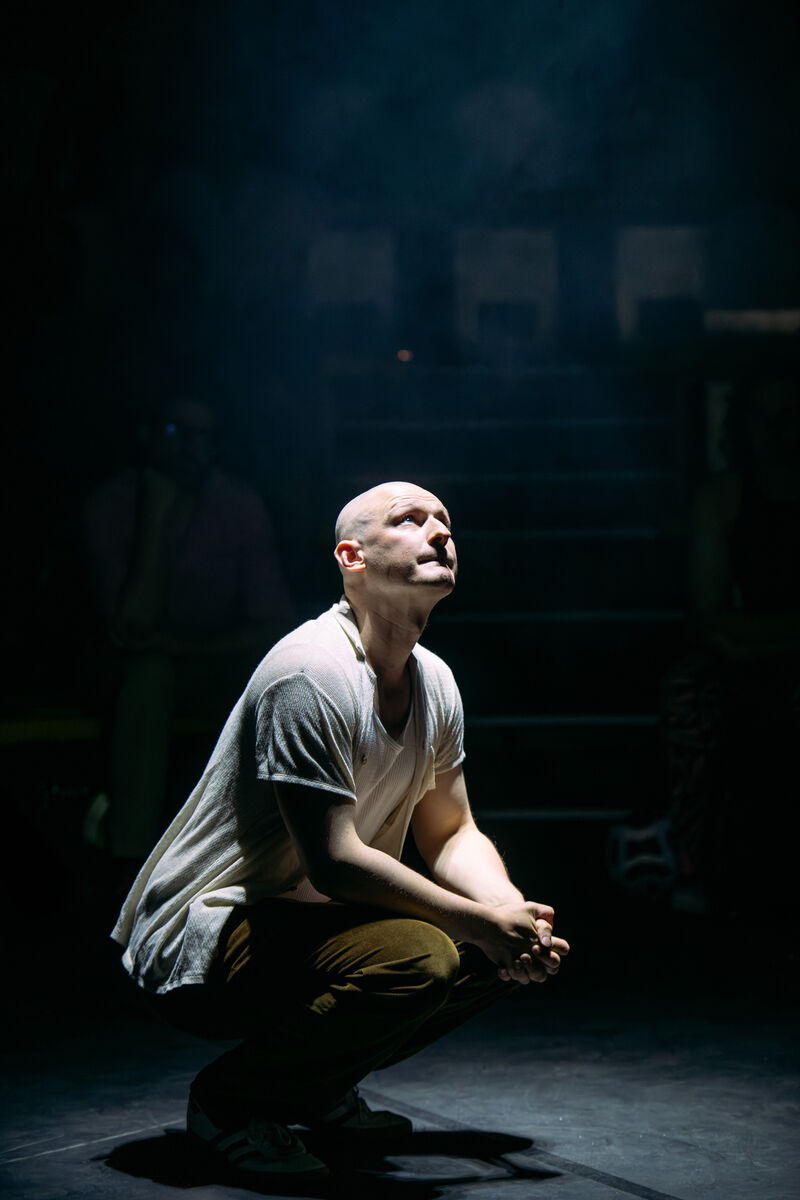 A white, bald, performer is crouching down and looking up at a stark white ligth