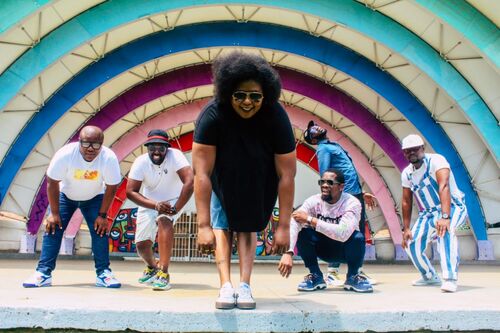 A black woman stands on a stage, she is bent over with her arms hanging at her side. Behind her stand 5 black men posing in various positions