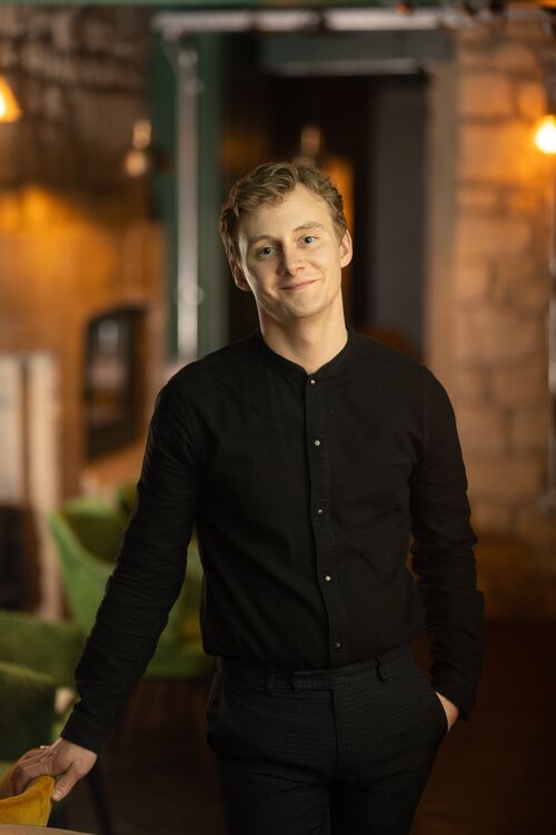 A young white man wearing a black shirt and trousers smiles towards the camera