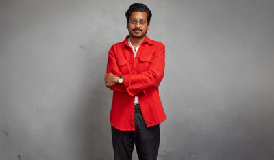 Ahir Shah wearing a bright orange shirt with his arms crossed over his chest against a grey background