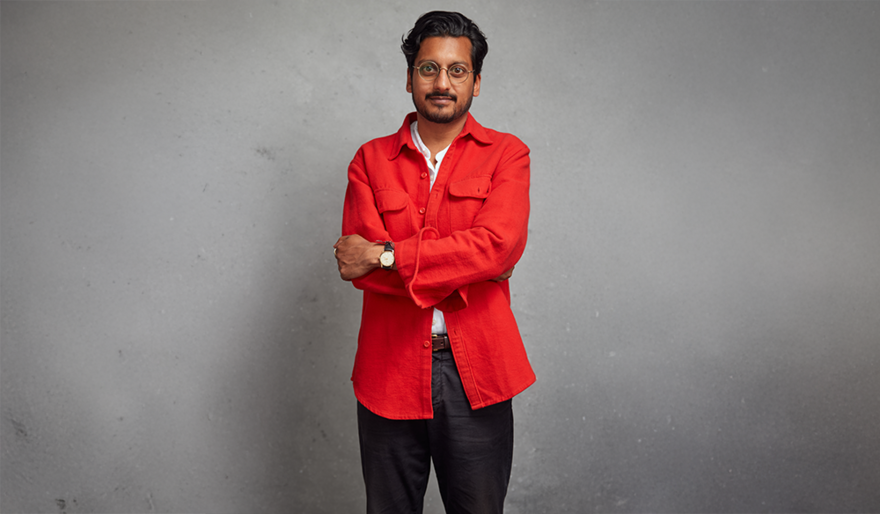 Ahir Shah wearing a bright orange shirt with his arms crossed over his chest against a grey background