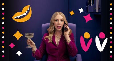 Katherine Ryan sitting on a purple chair holding a glass of champagne in one hand and a phone up to her ear. She is in a purple office with pink, yellow and white stars, dots and flowers surrounding her