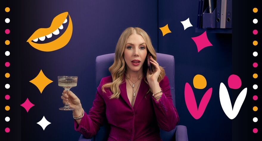 Katherine Ryan sitting on a purple chair holding a glass of champagne in one hand and a phone up to her ear. She is in a purple office with pink, yellow and white stars, dots and flowers surrounding her