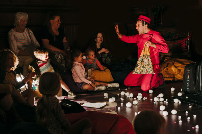  A performer in a red jumpsuit and hat is on their knees, interacting with a small group of adults and children sitting on the floor. The floor is covered in small, white, fluffy spheres. 