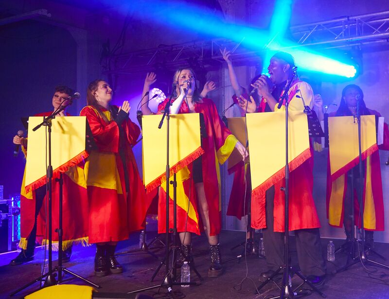 A choir stands singing into microphones on stage, they all have a pulpit and are wearing red and yellow robes