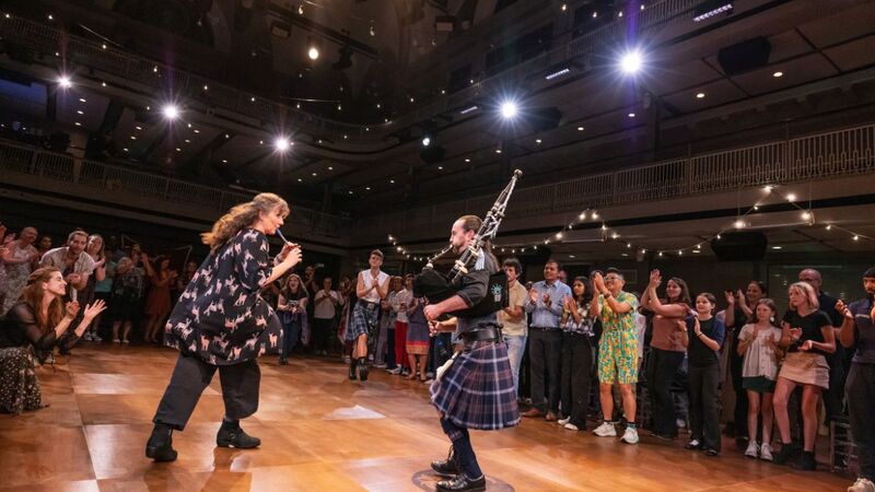 Two musicians face off on stage with string instrument, the audience is sat in the round around them