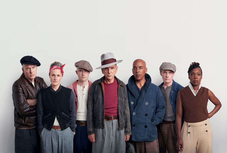 Group of 7 people standing in a line wearing varying hats. head scarfs and blazers