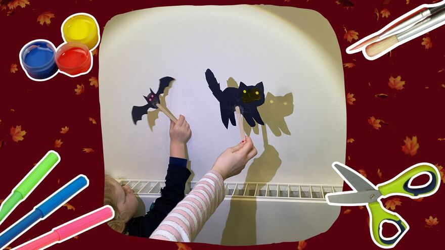 Two children holding up a bat and cat puppet, which is casting a shadow on the wall. This photo is on a red background with autumn leaves, and paints, brushes, pens and scissors.