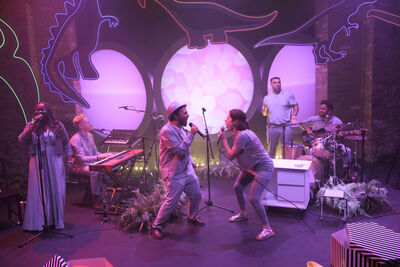 5 people on a purple stage with neon dinosaurs. There are two people facing each other singing, and others playing drums and keyboard. 