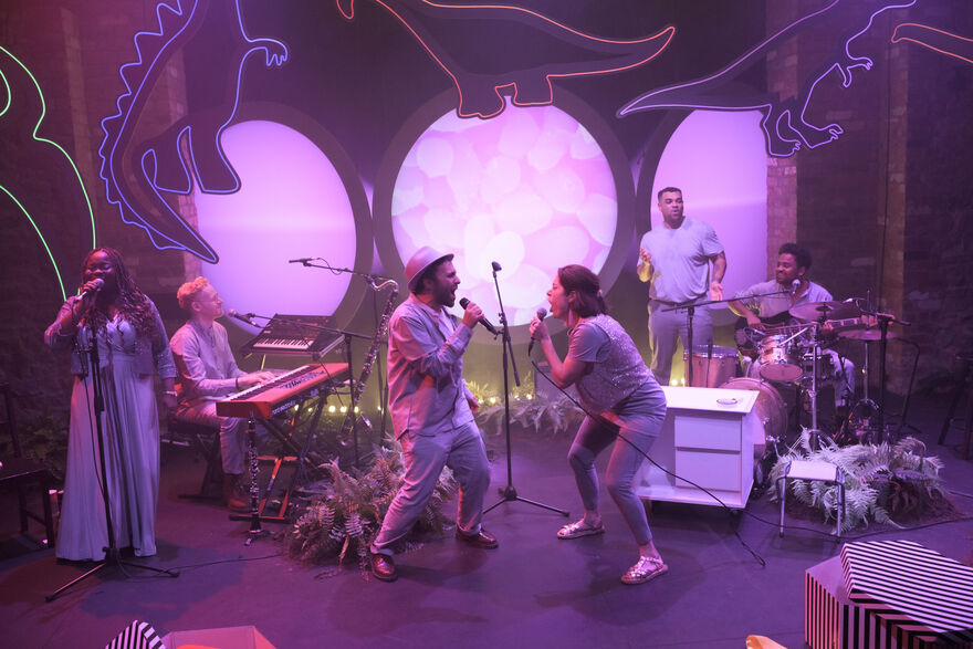 5 people on a purple stage with neon dinosaurs. There are two people facing each other singing, and others playing drums and keyboard. 