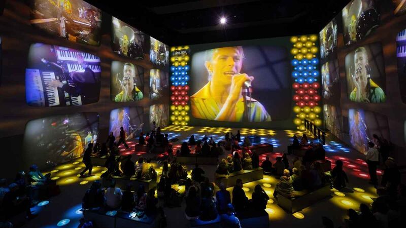 A dark immersive exhibition space with large screens showing a male singer performing, surrounded by colorful stage lights and seated spectators.
