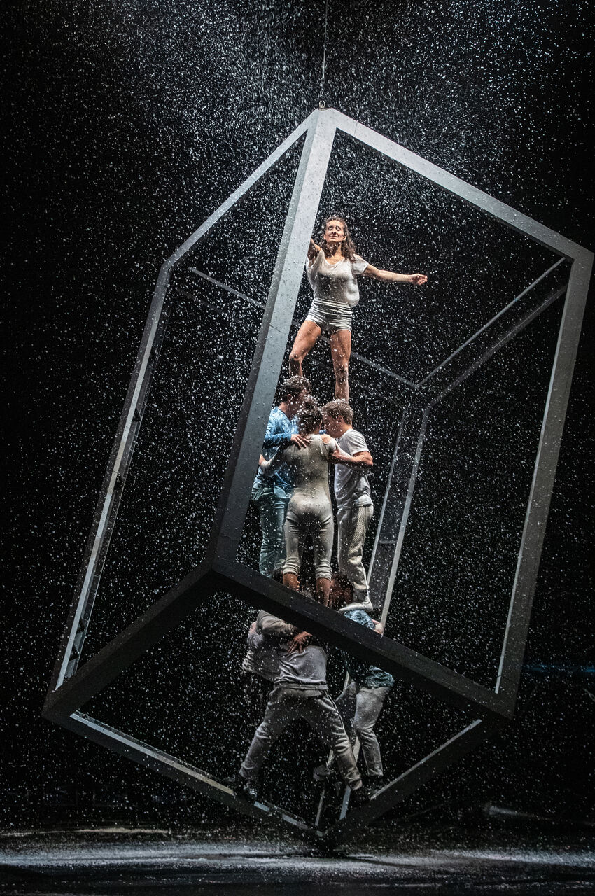 Six people performing a circus act by balancing within a cuboid shape with snow falling. There are three people at the bottom, holding up a further three, who are holding up a woman with her arms up.  