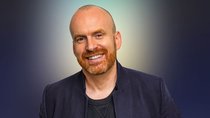 Image of author Matt Haig  who is smiling and wearing a black tshirt and blue jacket
