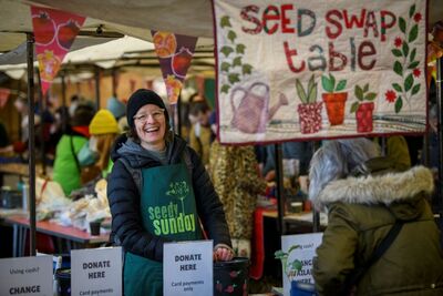 A person smiling standing behind a seed stall. Behind them is a sign which says 'Seed Swap Table'