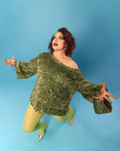 Jessica Fostekew is a green knitted jumper holding her arms out over a blue backgroun