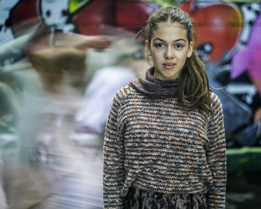 A young girl in a striped jumper stands in front of a wall covered in graffiti, behind her the are white blurrs