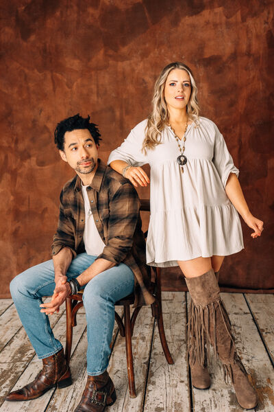 A man sitting on a wooden shirt, wearing a plaid shirt and jeans, a woman is leaning on the back of the chair