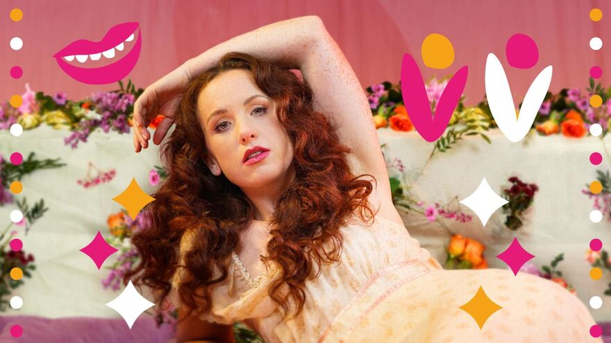 Catherine Bohart leaning down with her arm over her head in a creame dress, leaning on a purple mesh sheet and pink, orange purple flowers around her