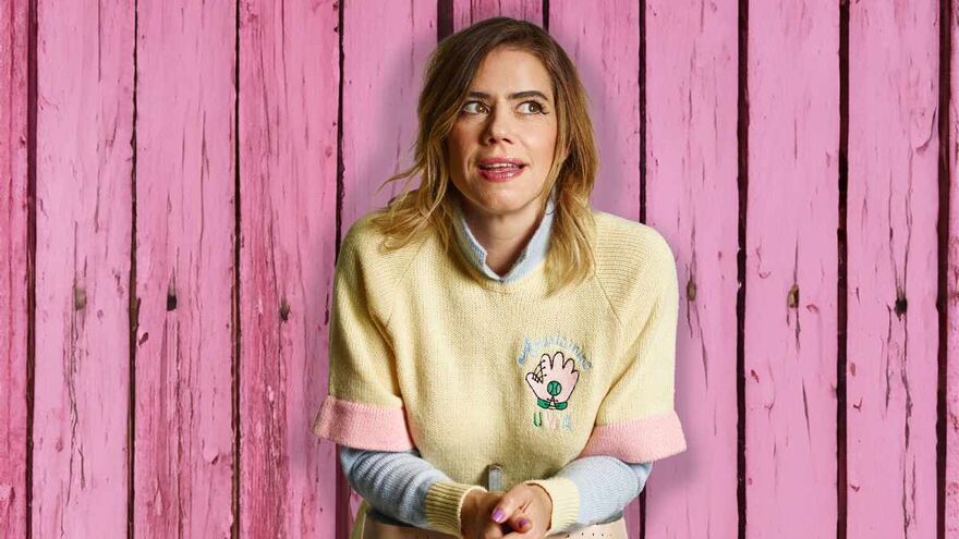 Lou Sanders standing against a pink wall wearing a playful yellow jumper. She is looking to the side of camera as if in the middle of speaking