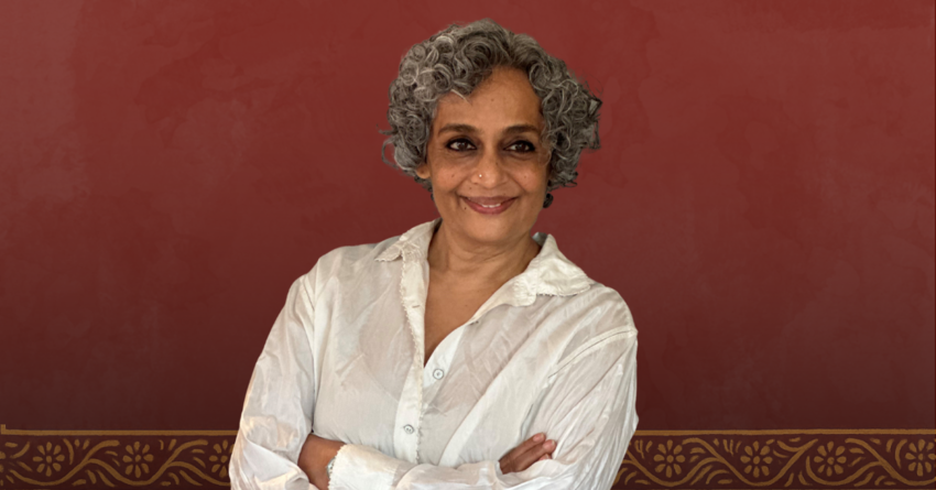 Arundhati Roy standing with her arms crossed over a red background