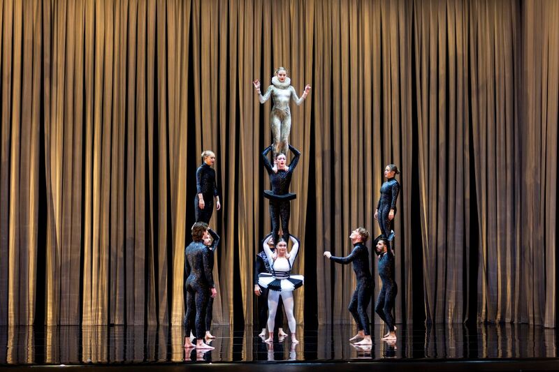 Eight people standing on a stage. Three people are standing on top of each others shoulders, either side are two people standing on each others shoulders