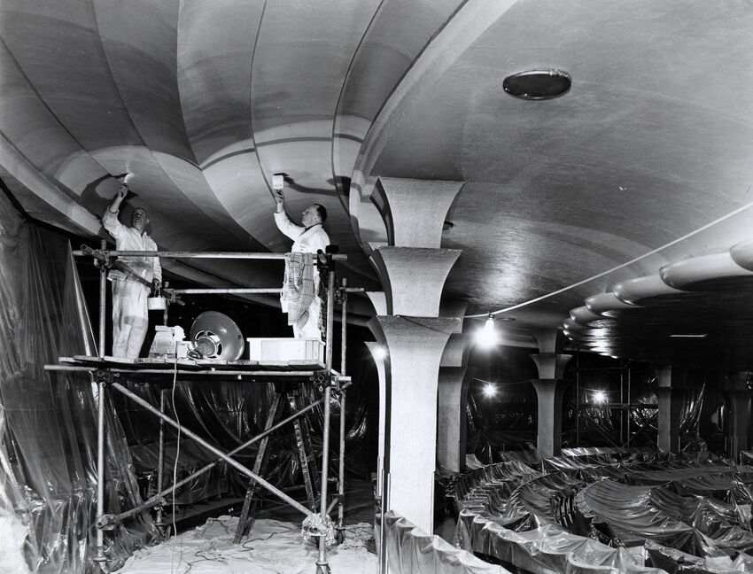 Black and white image of construction taking place within the Brighton Dome concert hall. Two men are standing on scaffolding, painting the ceiling