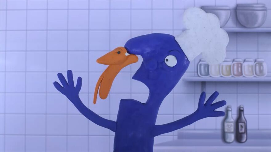A blue clay-style animated figure wearing a chef hat stands in a kitchen with tiled walls and shelves, with an orange clay animal attached to its nose with both arms raised