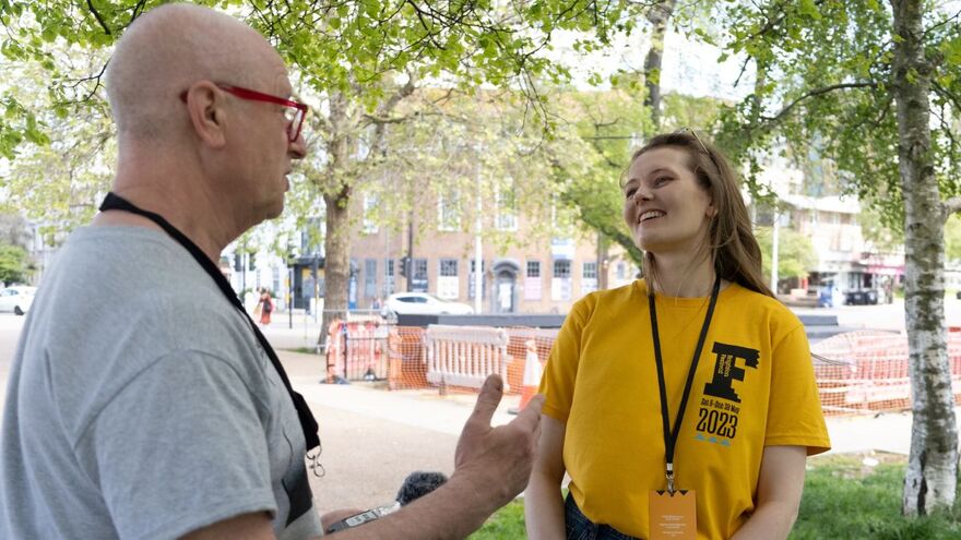 Two people stand speaking to one another. One is wearing a yellow t-shirt with a large Festival logo on it, and the other is wearing a grey t-shirt and red framed glasses