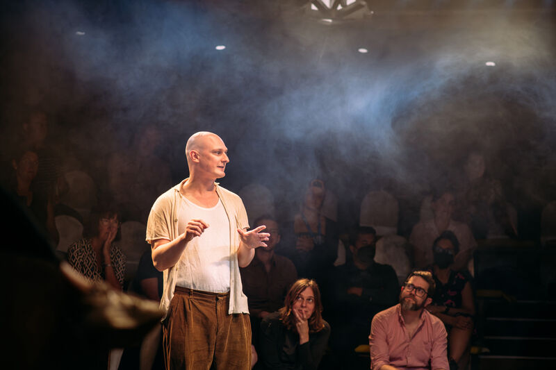 The performer stands in the middle of an in-the-round stage, with haze and ambient white lighting. He is white and bald, and the audience is sitting around him looking at his face