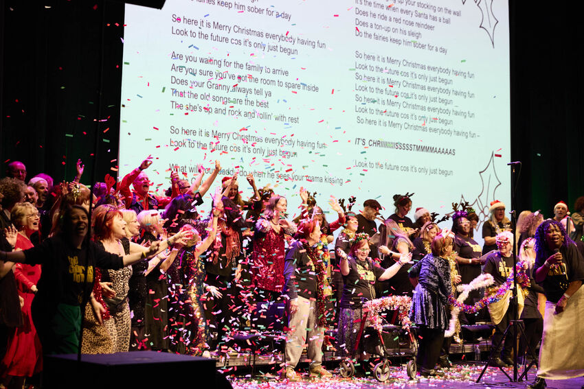 Loads of people celebrating on stage with confetti. They are wearing Christmads outfits including Reindeer antlers and tinsel. They are standing in front of a white background with Christmas song lyrics on it.