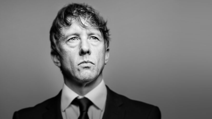 Jonathan Pie wearing a black tie and suit. The image is in black and white