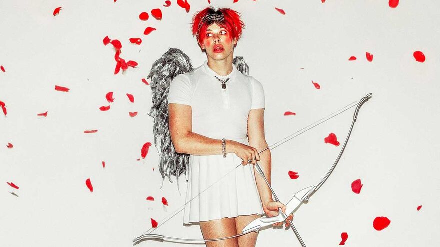 Yungblud with red hair and lipstick wearing a white dress with silver wings and a bow and arrow, surrounded by falling red petals