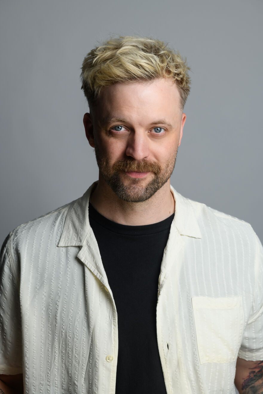 A man with blond hair and a beard staring at the camera. He is wearing a black top and a white short-sleeve shirt
